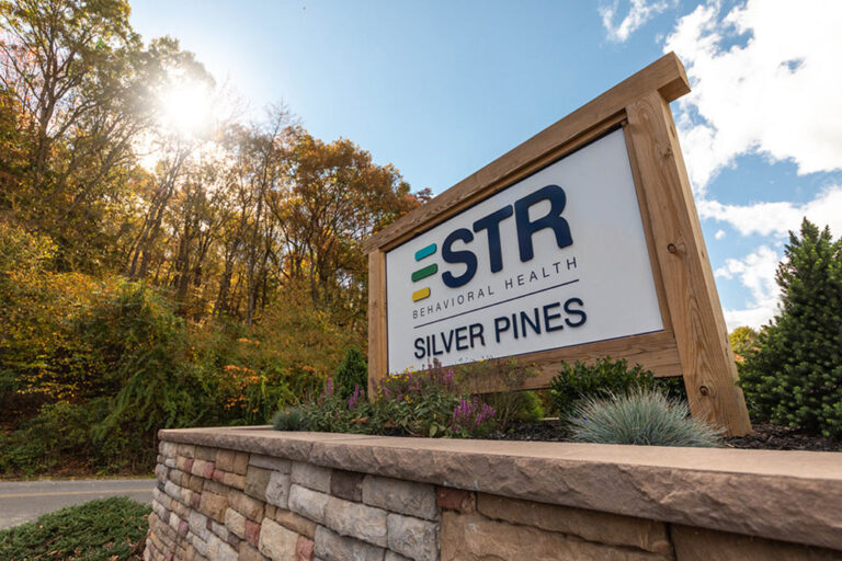 STR Behavioral Health Silver Pines (an addiction and mental health treatment center) signage against a sunny sky and wooded background.