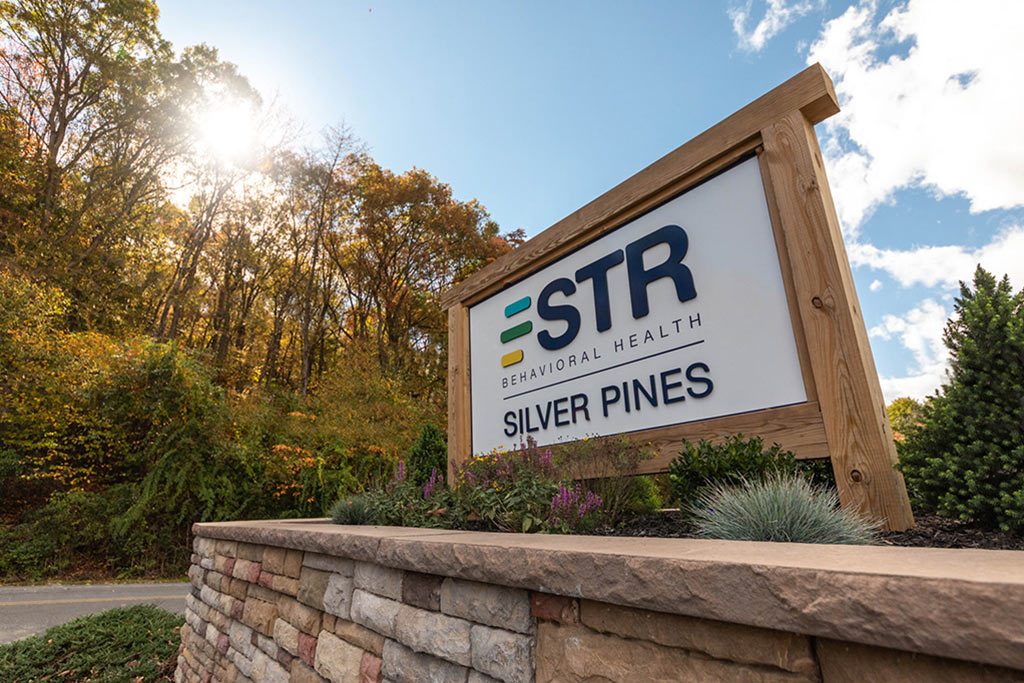 STR Behavioral Health Silver Pines (an addiction and mental health treatment center) signage against a sunny sky and wooded background.