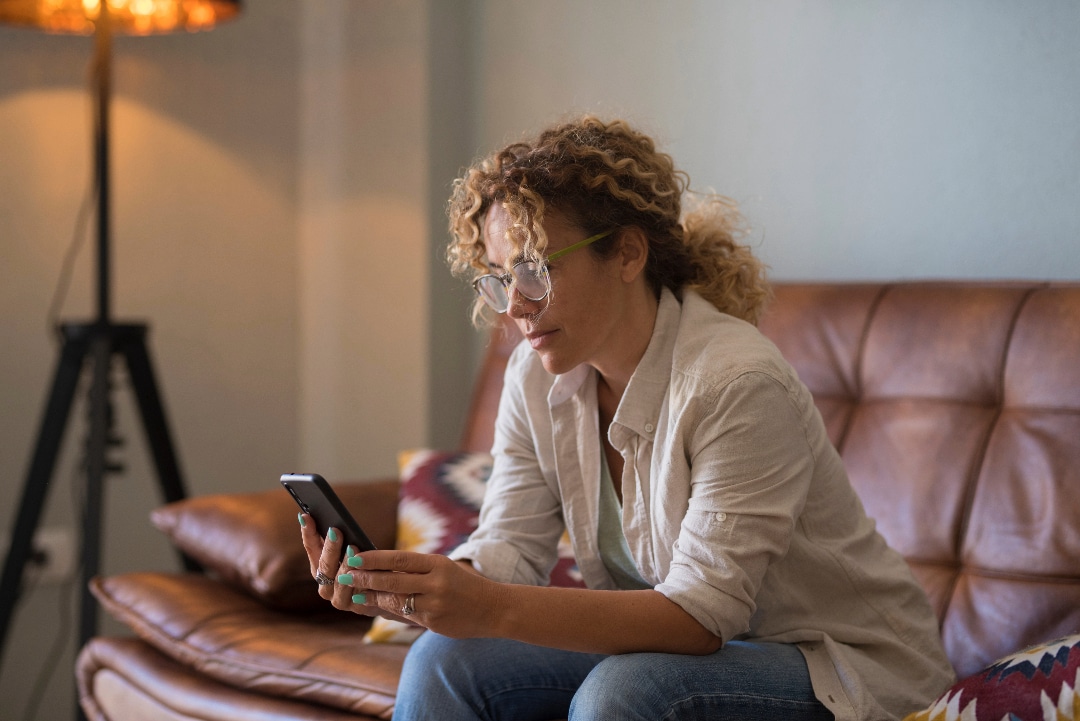 Woman sitting on her couch looking at her phone, contemplating whether to call a treatment center.