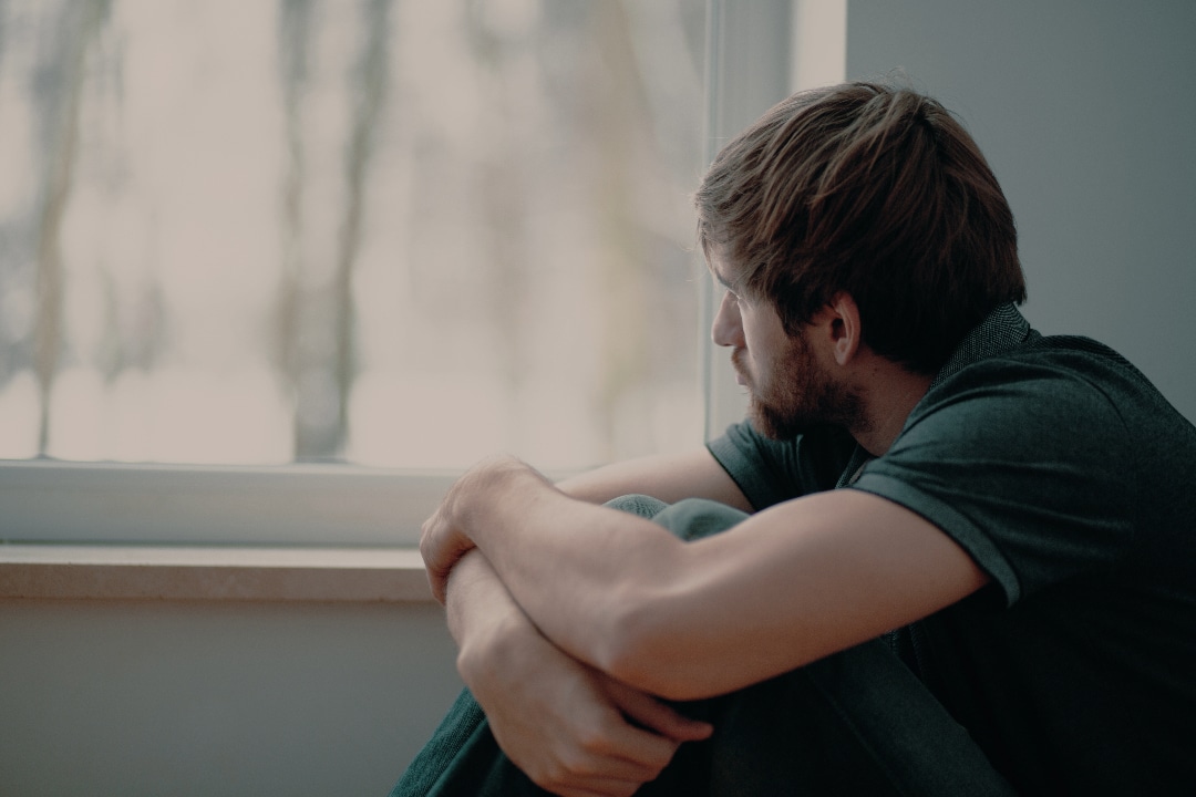 Young man sitting and looking out a window sadly.
